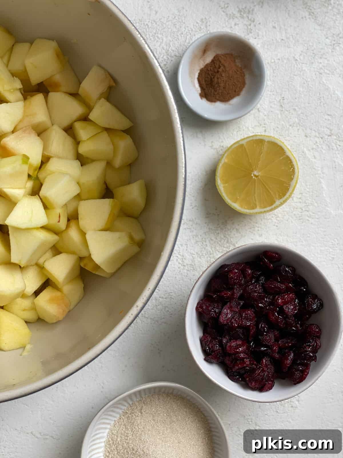 Autumn Apple Cranberry Delight 7 Ingredients for Apple Cranberry Crisp filling measured out against a white surface, including chopped apples, dried cranberries, cinnamon, and lemon juice.