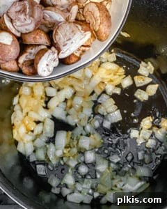 Sliced mushrooms being added to a black skillet containing sautéed onions and garlic.
