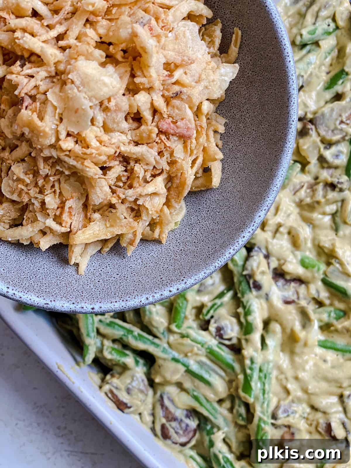 Plant-Powered Green Bean Casserole 7 Process shot of creamy green bean casserole being topped with a generous layer of crispy fried onions in a baking dish before baking.