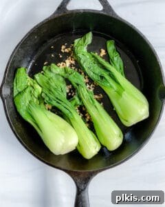 Garlic Soy Glazed Baby Bok Choy 6 The process of stir-frying: fragrant minced garlic sizzling in oil in a hot skillet, with blanched baby bok choy just added, ready to be tossed.