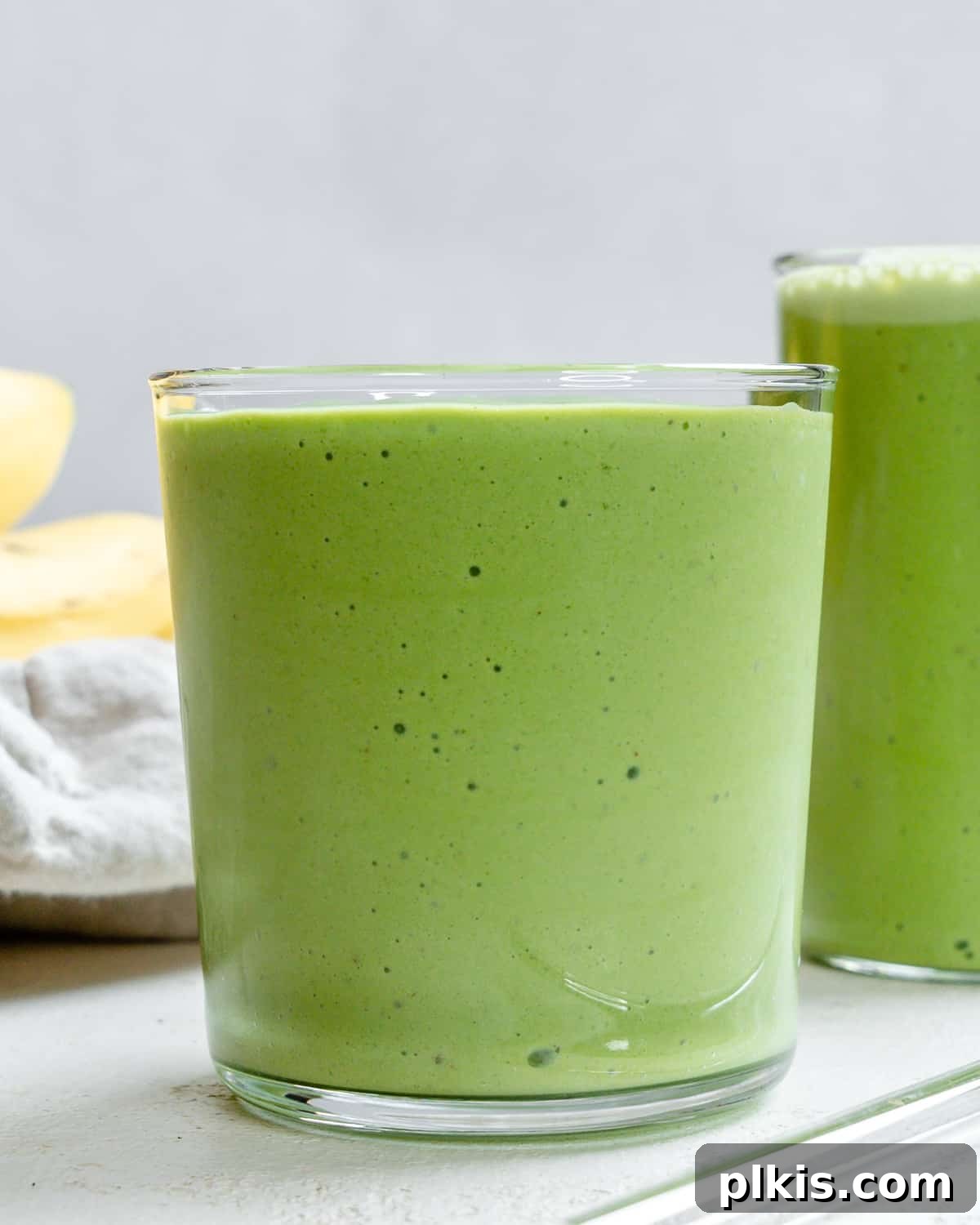 Creamy Kale Banana Bliss 2 Two glasses of vibrant green banana kale smoothie, garnished with banana slices and kale leaves, on a light background.