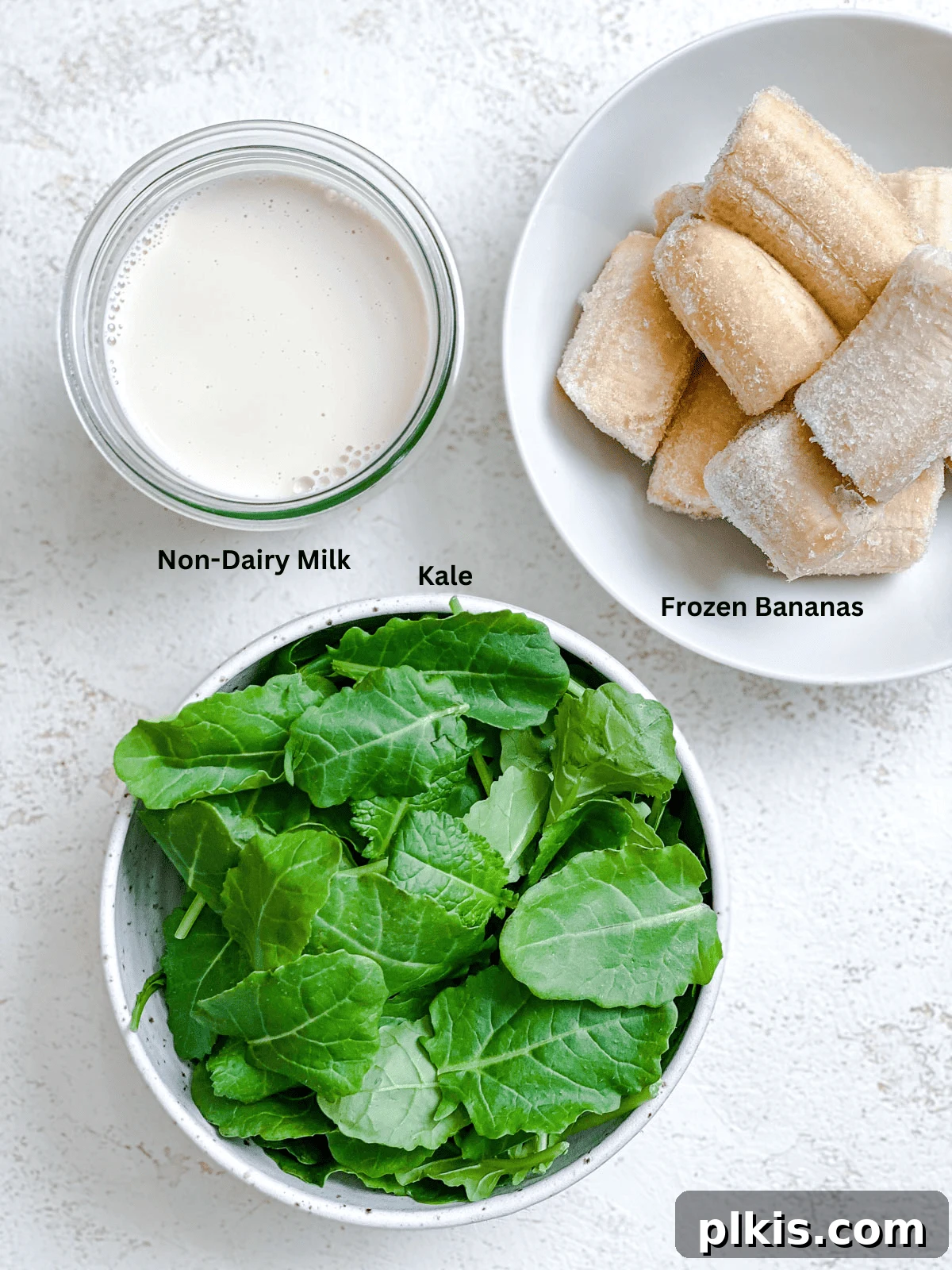 Creamy Kale Banana Bliss 3 Ingredients for a Banana Kale Smoothie measured out on a white surface, including frozen banana slices, fresh kale, and a glass of plant-based milk.