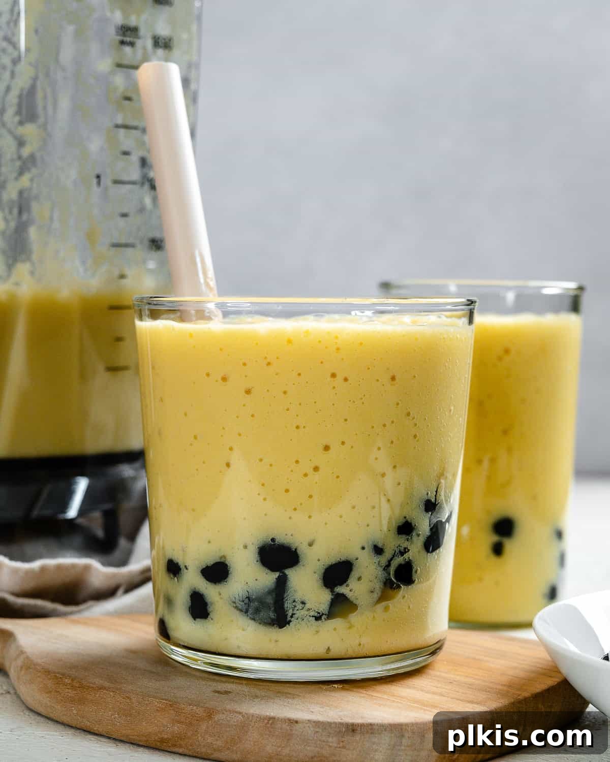 Tropical Mango Bubble 2 A tall glass of vibrant yellow Mango Bubble Tea with plump tapioca pearls at the bottom, topped with a frothy layer. Another similar glass is softly blurred in the background, creating an inviting and refreshing scene.