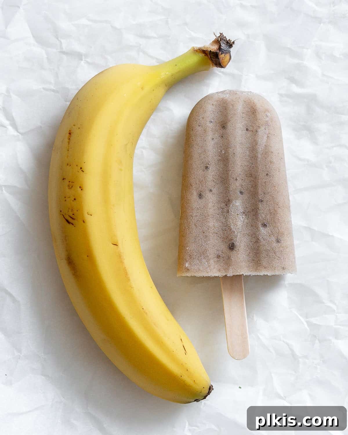 Naturally Sweet Banana Pops 7 A freshly made healthy banana popsicle alongside a whole banana on a pristine white surface, highlighting its natural goodness.