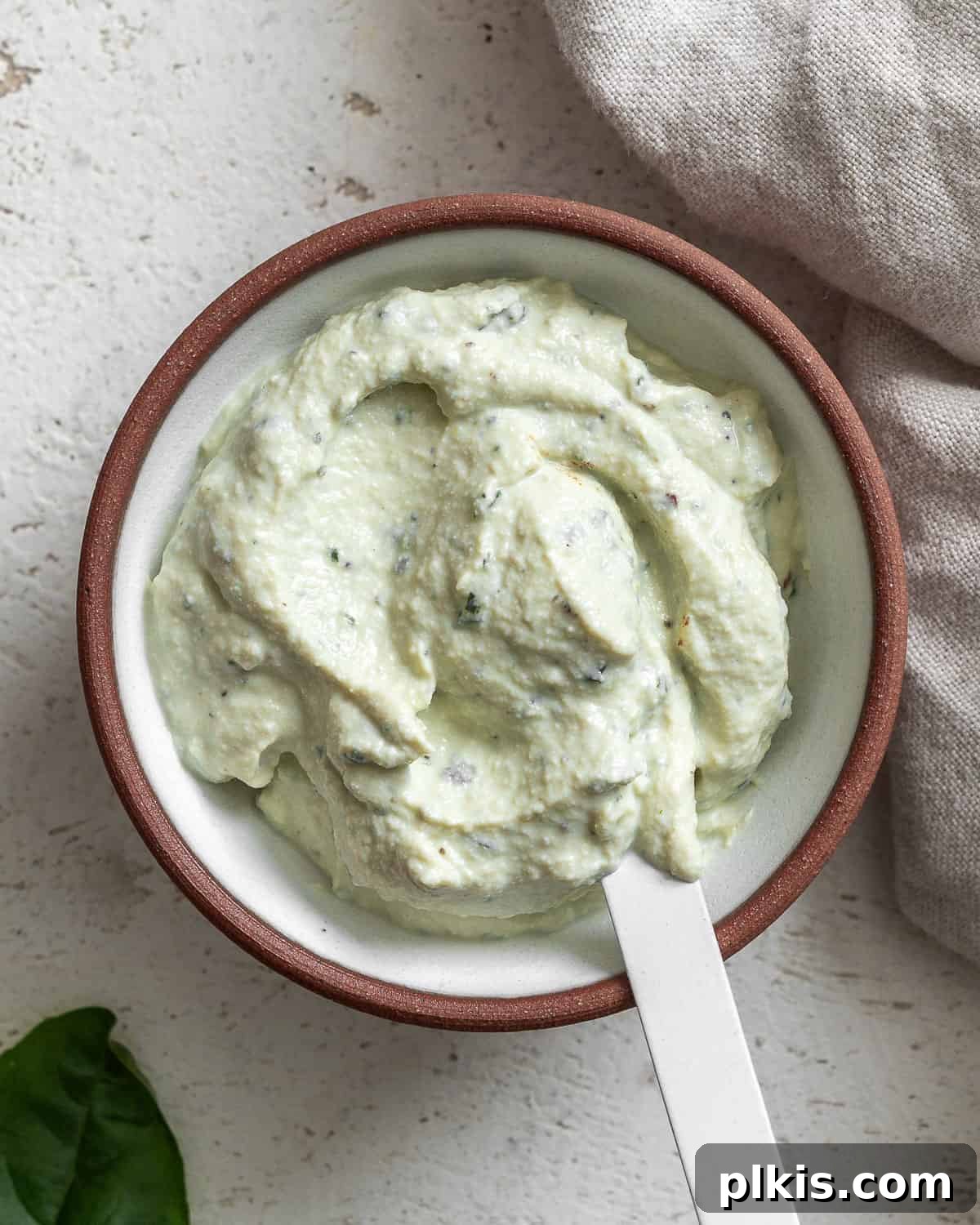 Creamy Vegan Tofu Ricotta 2 Bowl of tofu ricotta on a textured white surface with a white spoon in it and a leaf of basil in the bottom left corner.