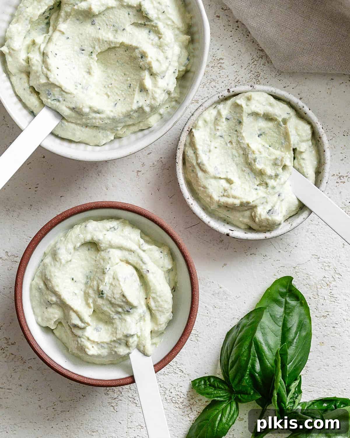 Creamy Vegan Tofu Ricotta 4 Three small bowls of tofu ricotta on white textured surface. Each bowl has a white spoon and there's a small bunch of fresh basil at the bottom right corner.