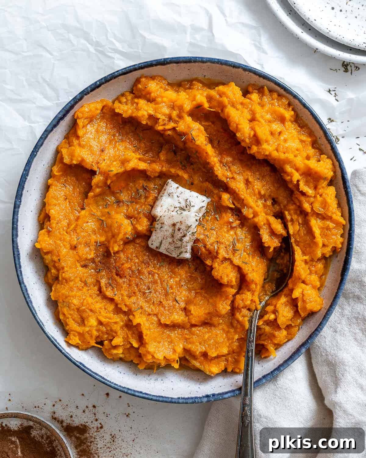 Velvety Vegan Sweet Potato Mash 2 Cooked mashed vegan sweet potatoes with a slice of vegan butter on top, served in a white bowl with a blue rim and a silver spoon. Photographed on crinkled white paper with a white cloth napkin and cinnamon in the foreground.