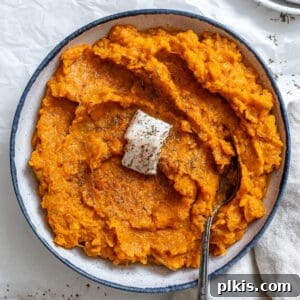 Cooked mashed vegan sweet potatoes with a slice of vegan butter on top, served in a white bowl with a blue rim and a silver spoon.