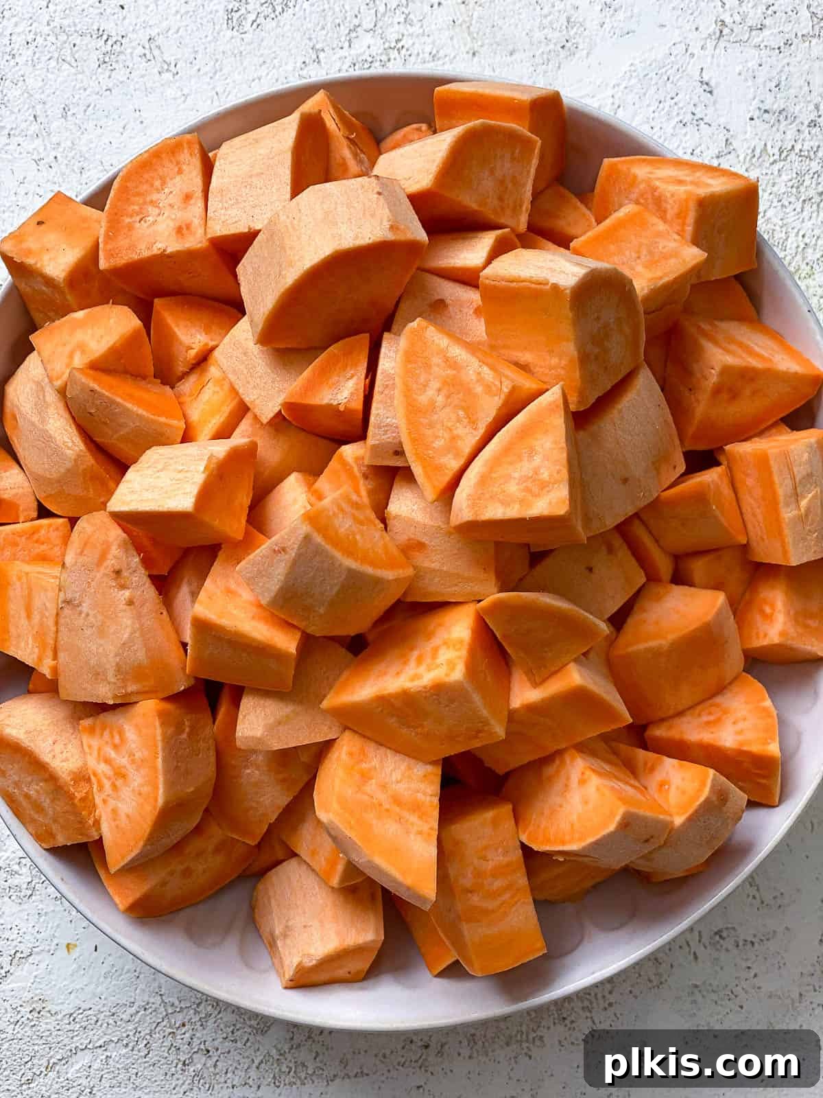 Velvety Vegan Sweet Potato Mash 4 A bowl of raw chopped and peeled sweet potatoes, ready for cooking, set on a textured white surface.