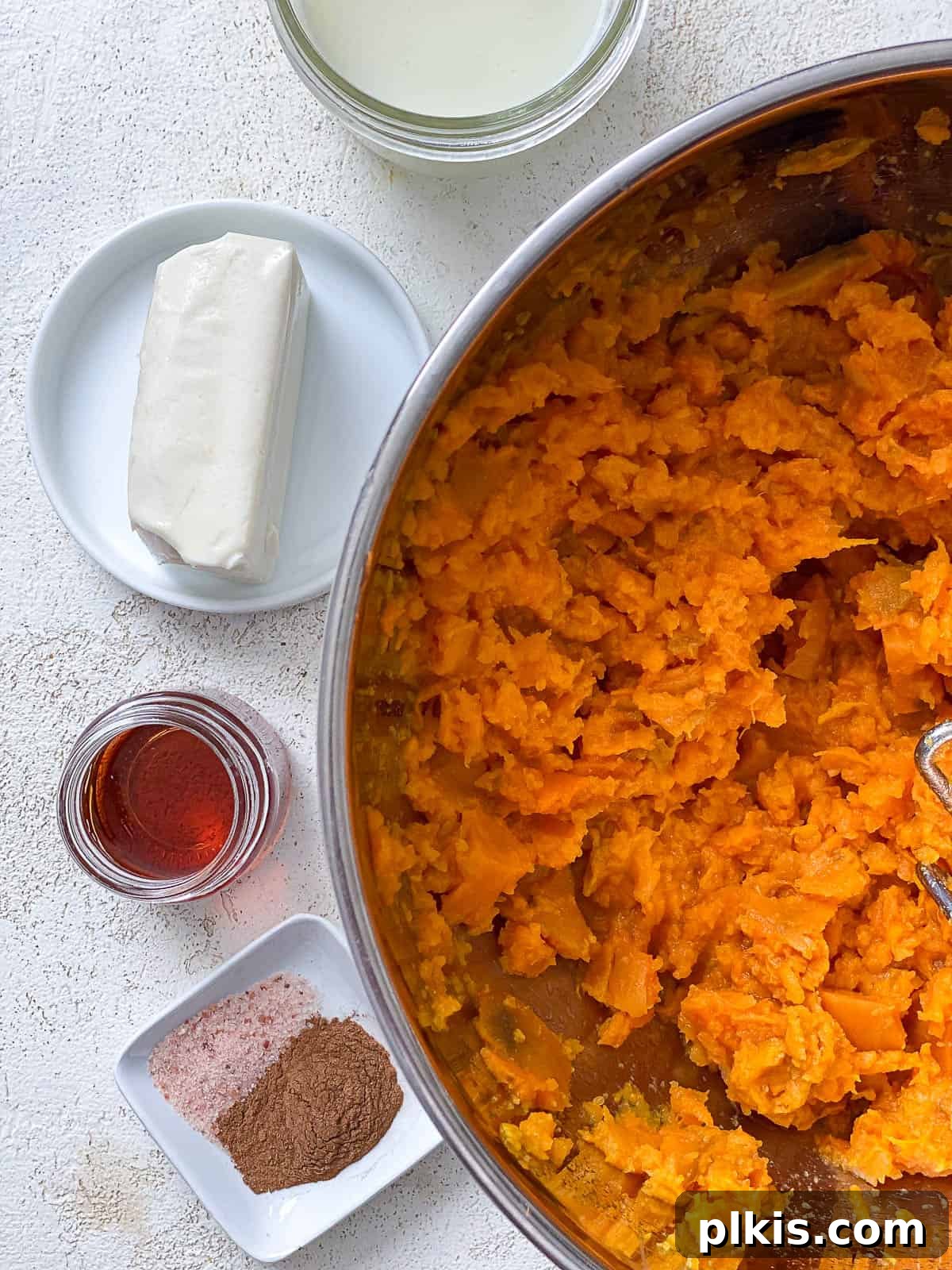 Velvety Vegan Sweet Potato Mash 8 Mashed sweet potatoes in a silver bowl, surrounded by small containers of vegan butter, plant milk, salt, cinnamon, and maple syrup, ready for final seasoning.
