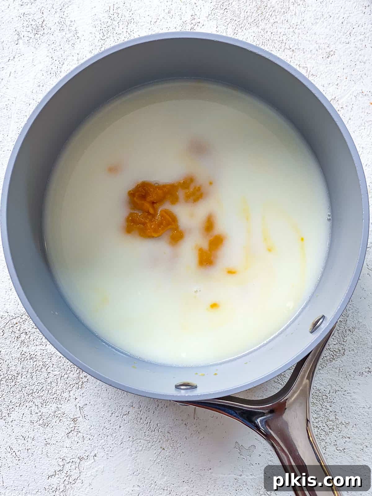 The Ultimate Plant-Based Pumpkin Spice Latte 4 A gray pot with a silver handle on a white textured surface, containing white milk and a dollop of orange pumpkin puree, ready to be mixed.