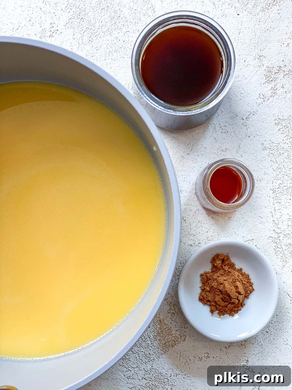 The Ultimate Plant-Based Pumpkin Spice Latte 5 A gray pot with milk and pumpkin puree gently mixed inside, sitting on a textured white surface. Beside it are small bowls containing sweetener, vanilla extract, and pumpkin pie spice, ready to be added to the mixture.