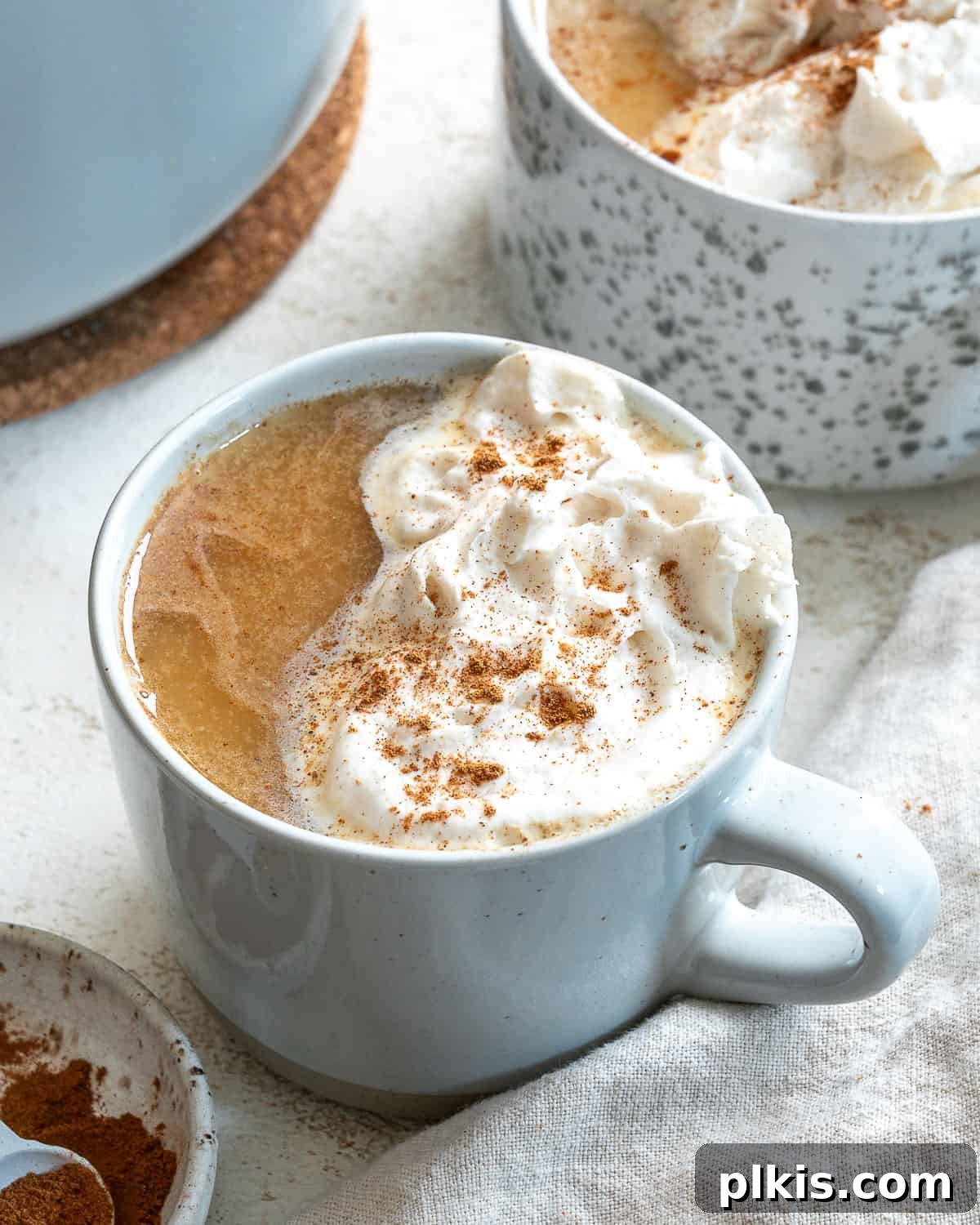 The Ultimate Plant-Based Pumpkin Spice Latte 9 A finished vegan pumpkin spice latte topped with vegan whipped cream. On the top left corner, theres part of another glass with the same drink. Bottom right has a gray napkin. Bottom left has a small bowl of cinnamon.