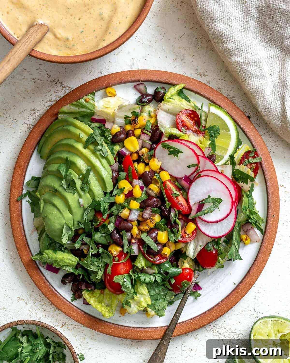 Vegan Fiesta Bowl 2 A vibrant vegan taco salad presented in a crispy tortilla bowl, generously topped with sliced avocado, a creamy taco salad dressing, fresh radishes, and bright lime wedges. In the background, a small bowl of dressing with a wooden spoon, a gray napkin, and a side arrangement of chopped cilantro, half an avocado, and sliced radishes complete the inviting scene against a textured white backdrop.