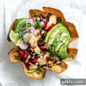 A tortilla bowl holding a taco salad that's topped with a sliced avocado, taco salad dressing, sliced radishes, and lime wedges