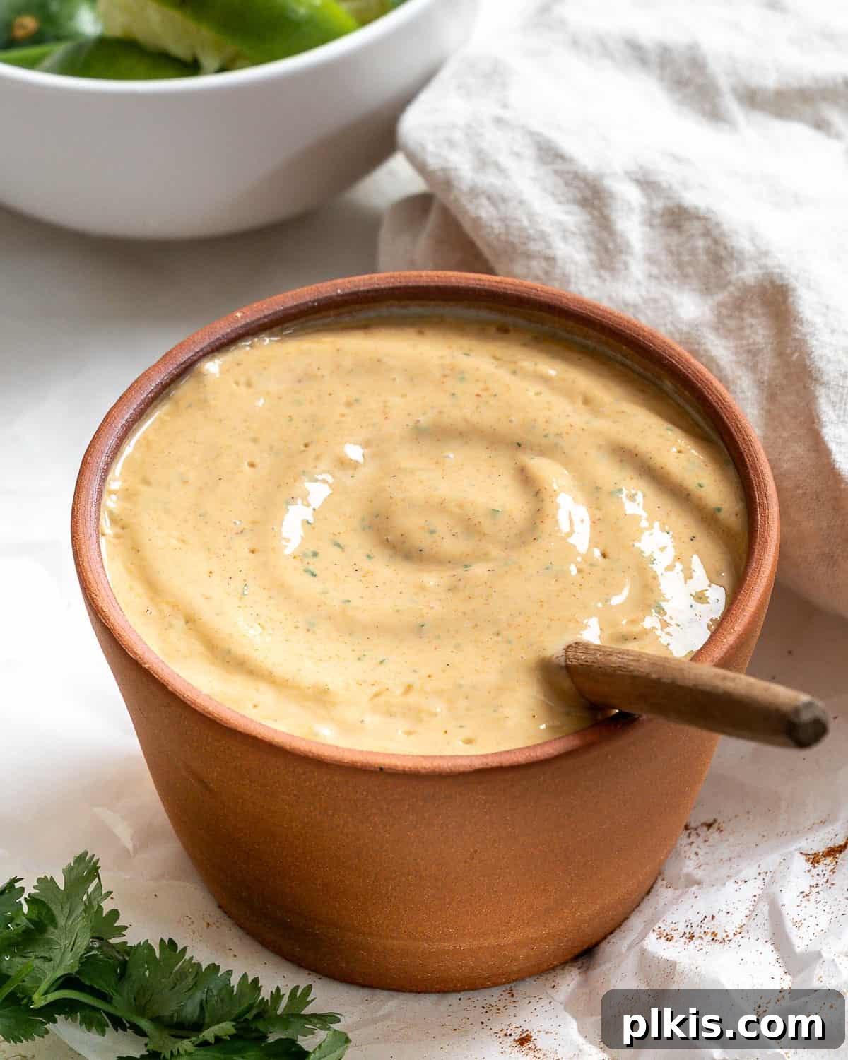 Vegan Fiesta Bowl 4 A small terracotta bowl filled with creamy taco salad dressing, accompanied by a wooden spoon. Fresh cilantro sprigs are visible in the bottom left, and a neatly folded gray napkin rests in the top right corner.