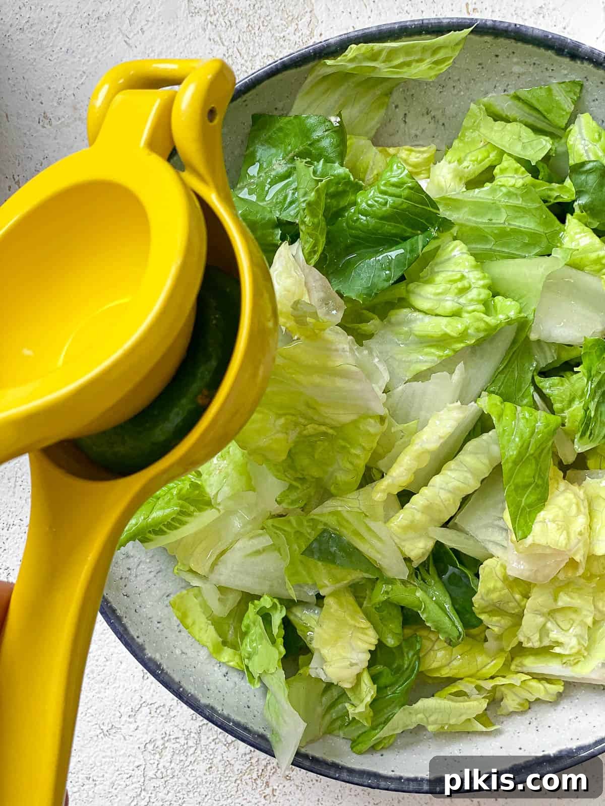 Vegan Fiesta Bowl 6 A white bowl with a subtle blue rim, filled with crisp chopped romaine lettuce. A yellow citrus squeezer is actively pressing lime juice into the bowl, adding a fresh zest.