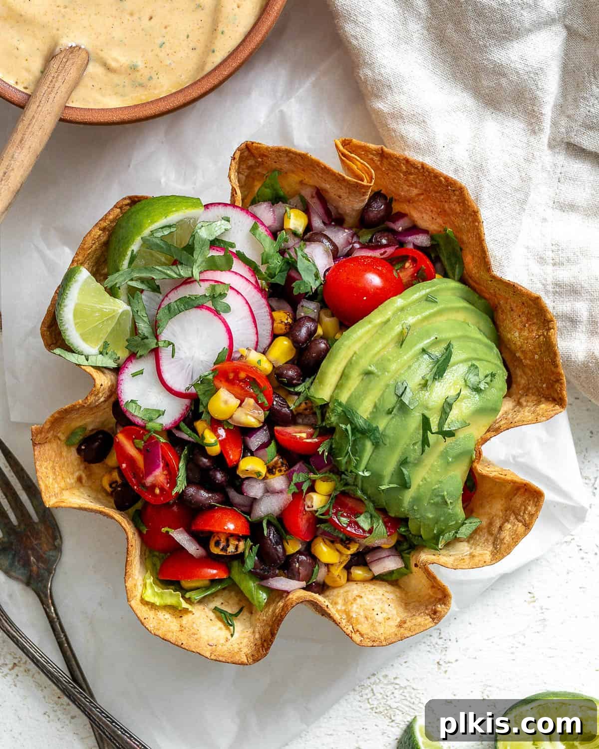 Vegan Fiesta Bowl 9 A meticulously prepared vegan taco salad nestled within a golden, crispy tortilla bowl. The salad is richly topped with creamy sliced avocado, a generous drizzle of savory taco salad dressing, thinly sliced radishes for crunch, and bright lime wedges for garnish. In the upper left corner, a small bowl holds extra dressing with a wooden spoon, while a gray napkin rests in the upper right. The lower left corner displays a small bowl of chopped cilantro alongside half an avocado and more sliced radishes, all set against a white, textured background.