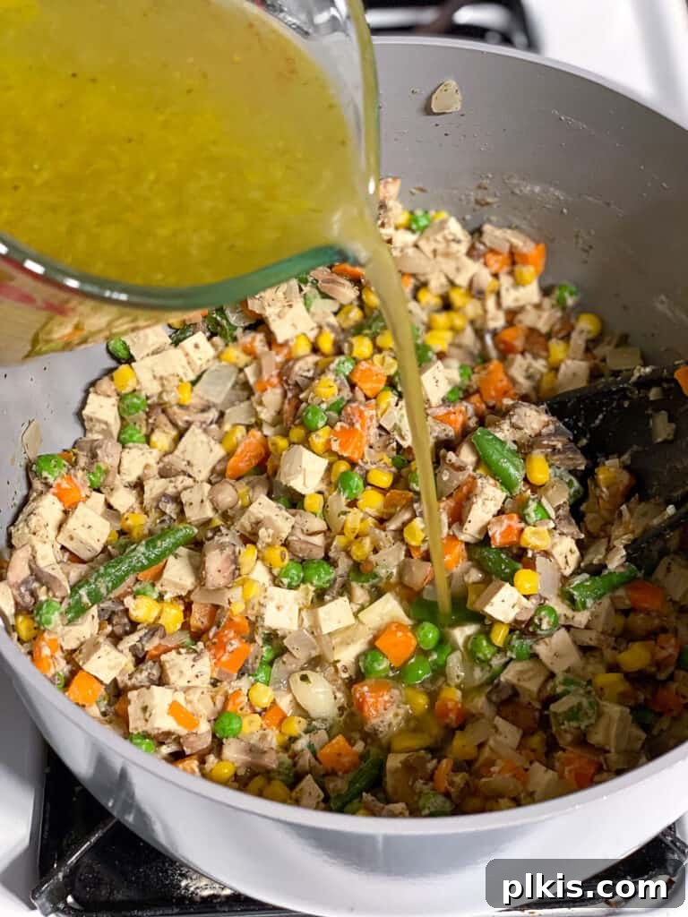 Vegetable broth is being slowly poured into the flour-coated vegetable mixture in the pan, forming the rich, savory sauce for the vegan pot pie.