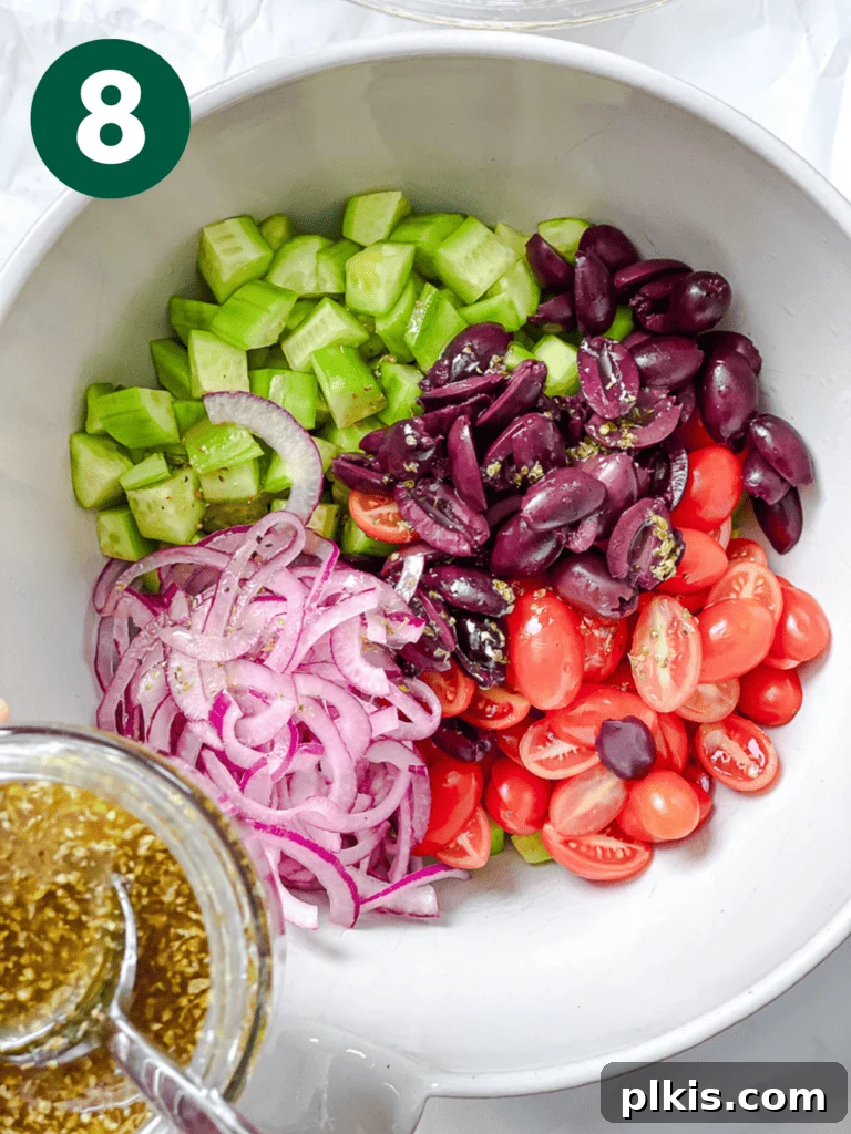Process shot demonstrating the Greek salad dressing being drizzled over the vibrant vegetable mixture in a large bowl.