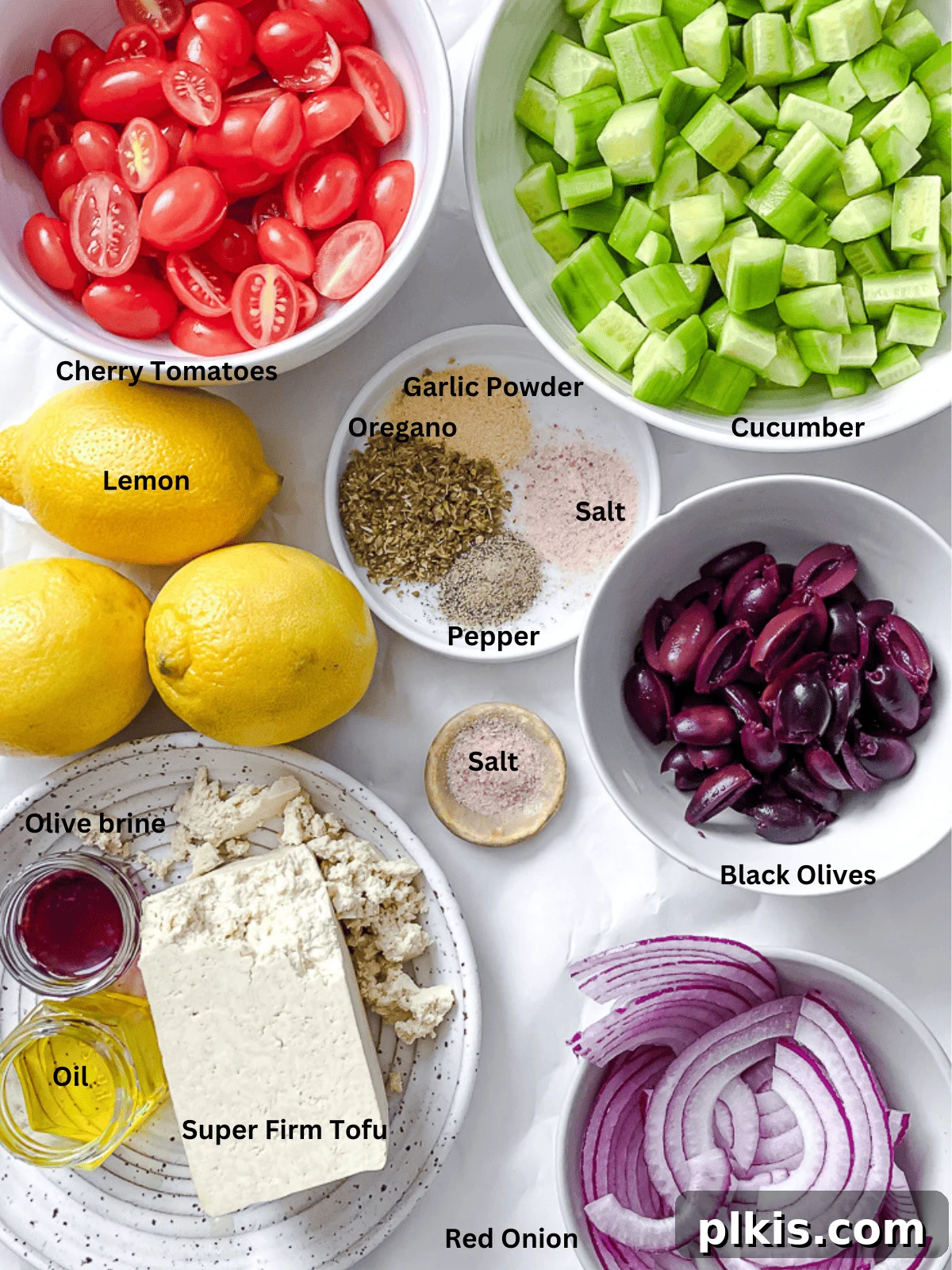 A pristine white surface displays an organized array of fresh ingredients for Greek Cucumber Salad, including crisp cucumbers, ripe tomatoes, black olives, red onion, lemon, and a block of tofu, ready for preparation.