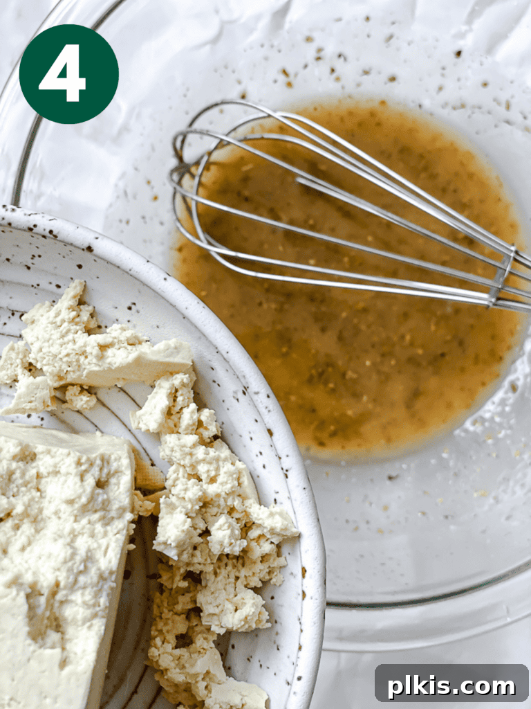 Process shot illustrating crumbled tofu being added to the whisked marinade in a bowl, ready to absorb the Greek flavors.
