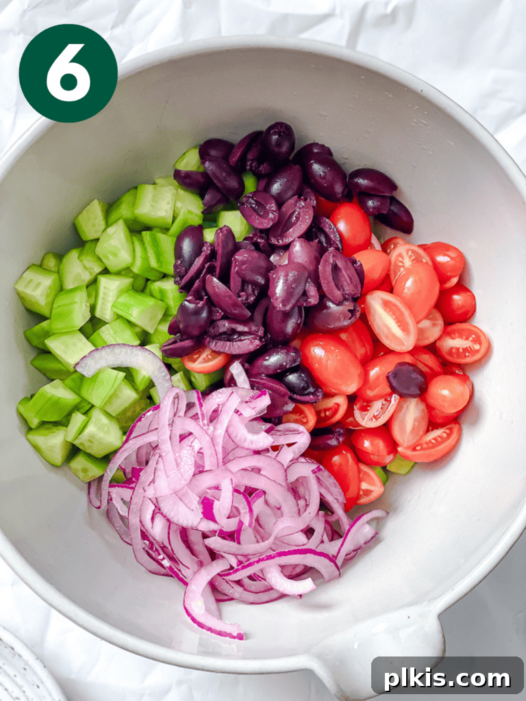 A large salad bowl filled with an colorful array of diced cucumbers, halved cherry tomatoes, sliced black olives, and drained red onions, ready for dressing.