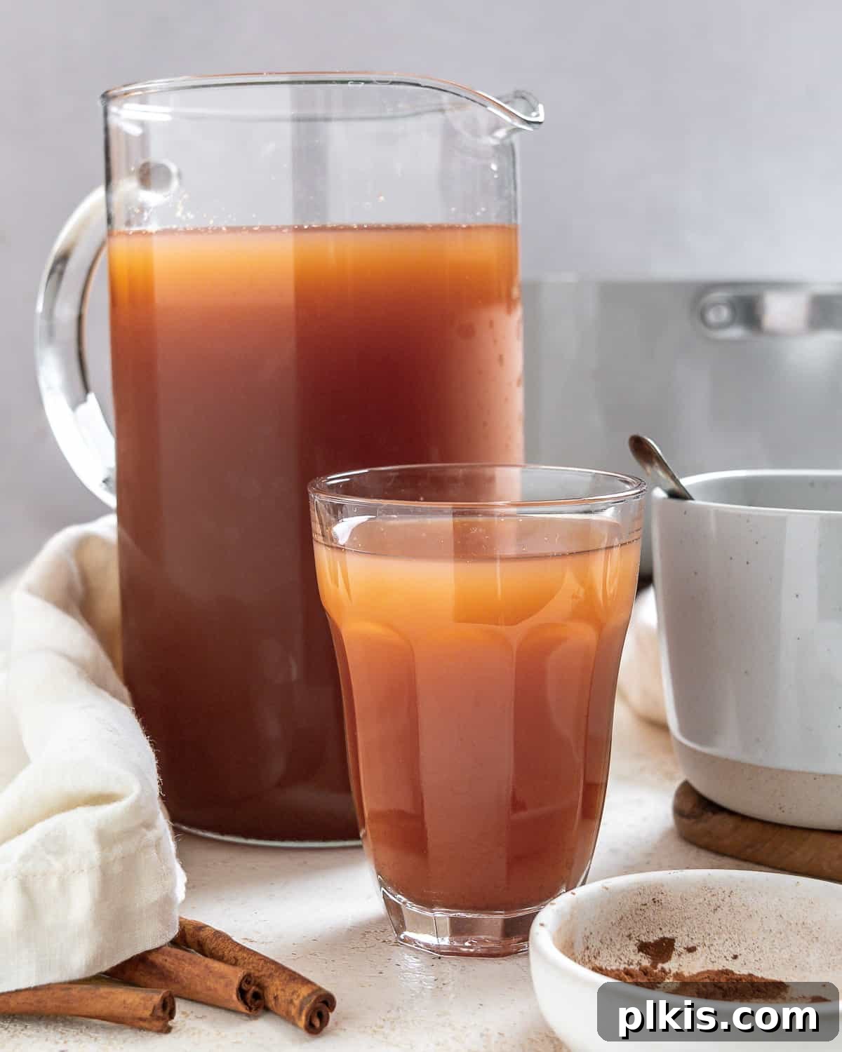 Cozy Spiced Apple Warmer 2 Two steaming cups of mulled apple cider next to a pitcher, garnished with cinnamon sticks and apple slices.