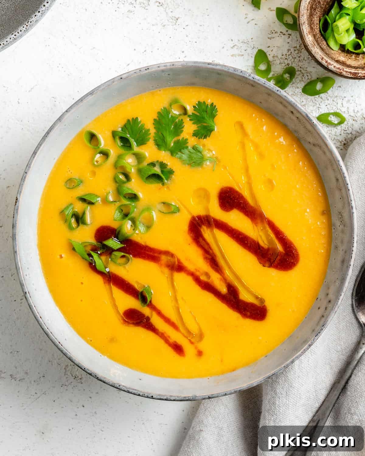 Golden Thai Squash Elixir 2 A beautifully presented bowl of Thai Butternut Squash Soup, garnished with fresh cilantro and a swirl of sriracha, set against a pristine white background. This comforting vegan soup offers a perfect balance of sweet, spicy, and creamy flavors.
