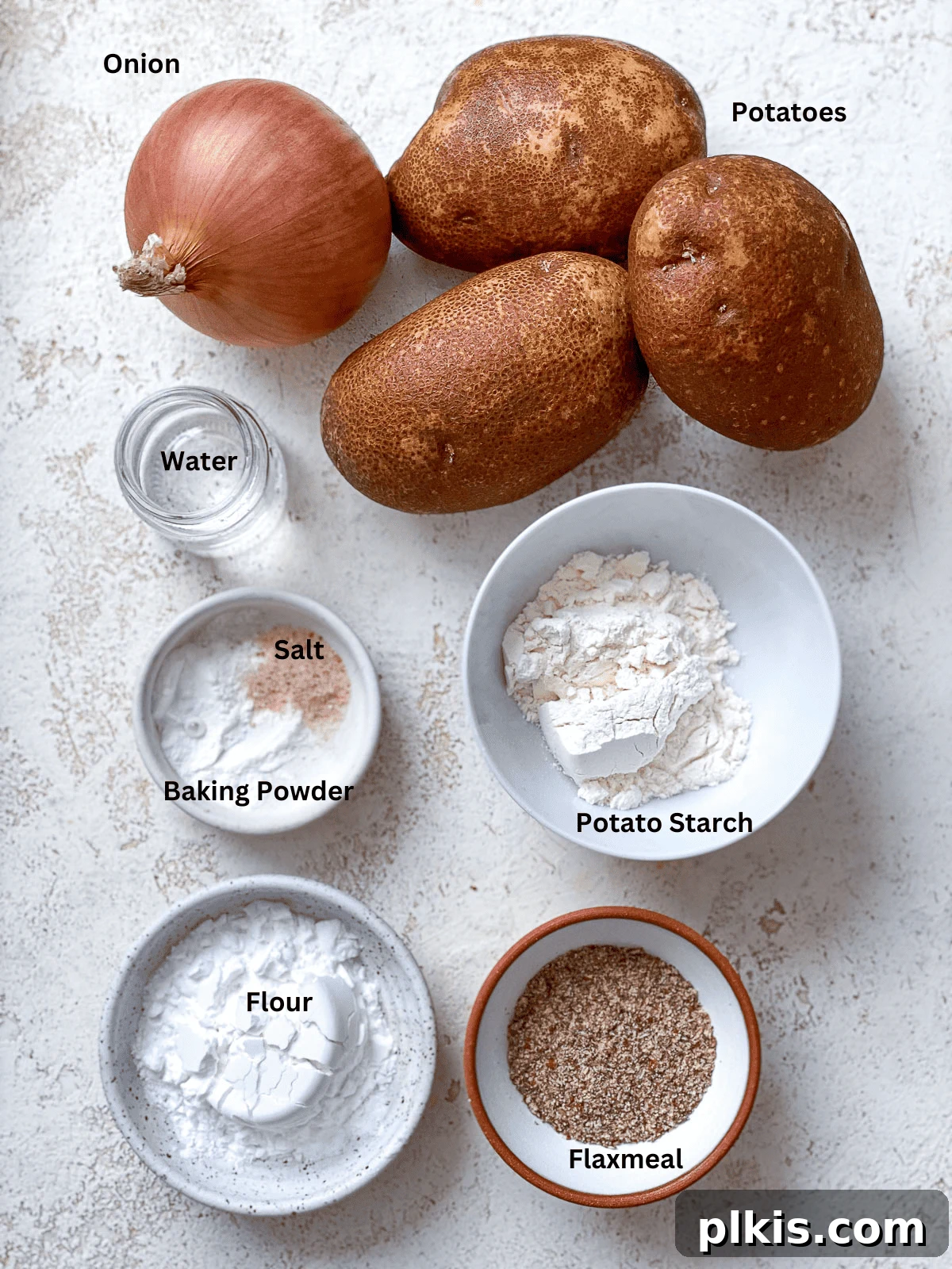 Crispy Vegan Potato Latkes 3 A vibrant collection of ingredients for Vegan Potato Latkes laid out on a white surface, including russet potatoes, an onion, flax meal, flour, and a bowl of spices, ready for preparation.