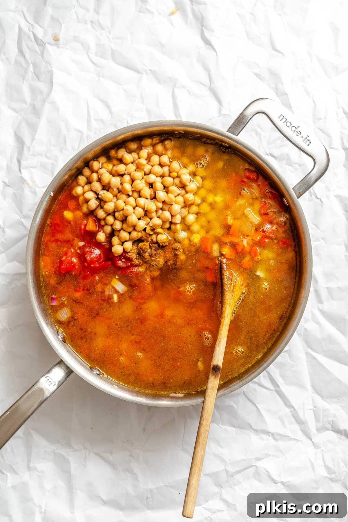 Process shot showing the vibrant mix of diced tomatoes, chickpeas, broth, and spices being stirred into the sautéed vegetables in the pot, simmering to infuse flavors.