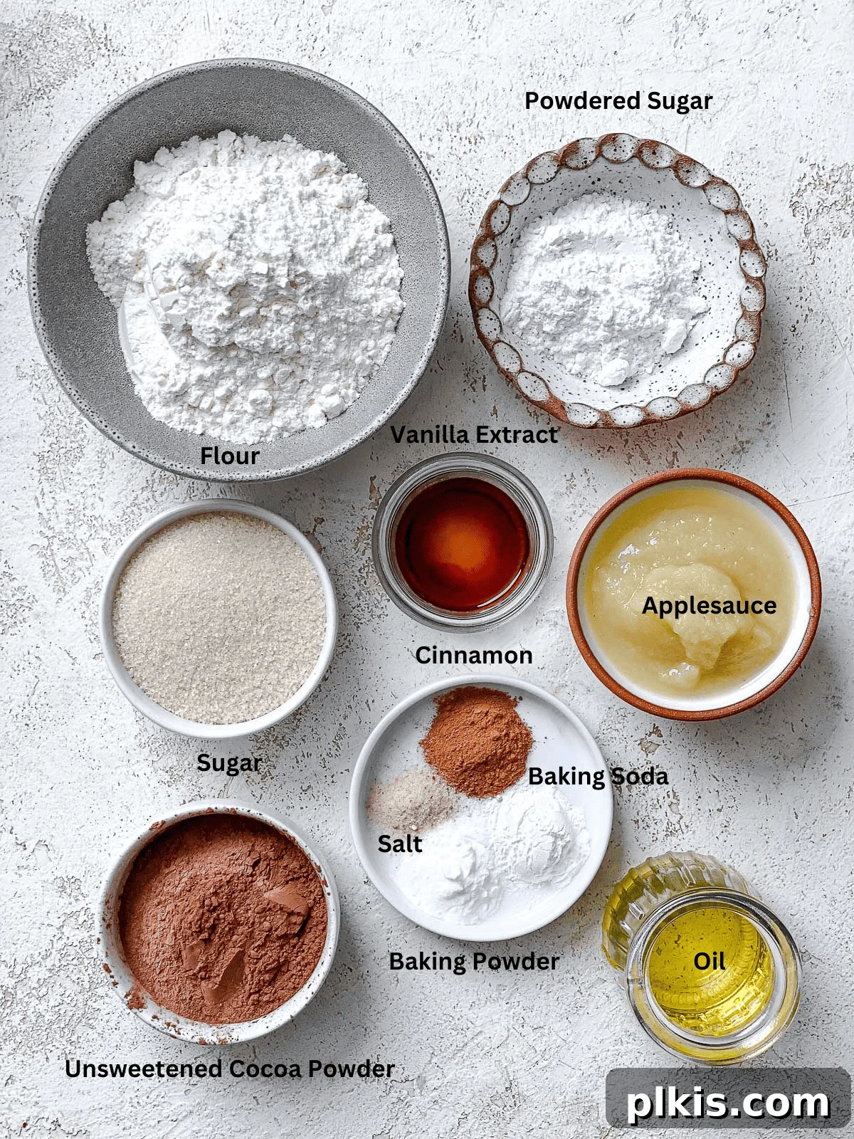 A collection of ingredients laid out on a white surface, including cocoa powder, sugar, oil, applesauce, flour, and spices, ready for making vegan chocolate crinkle cookies.