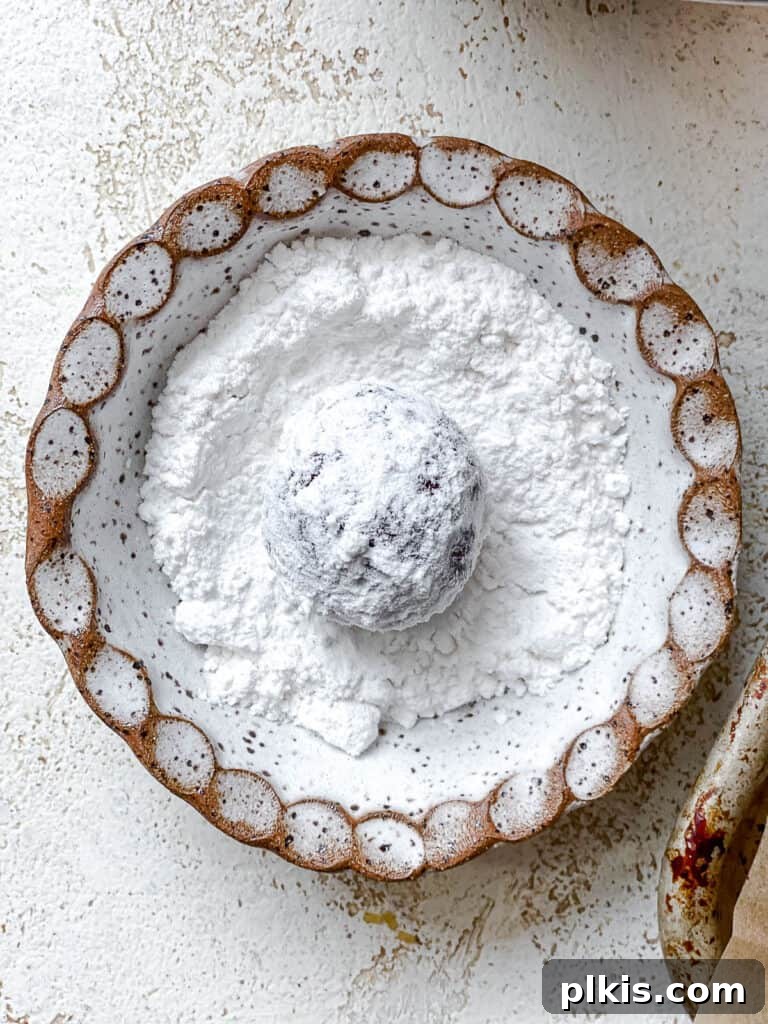 Process shot showing a ball of cookie dough being rolled in a bowl of powdered sugar, ensuring a thick coating.