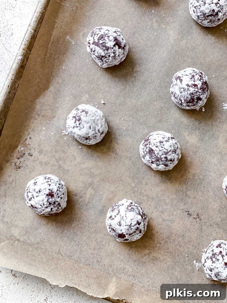Process shot displaying unbaked cookie dough balls heavily coated in powdered sugar, arranged on a baking sheet before going into the oven.