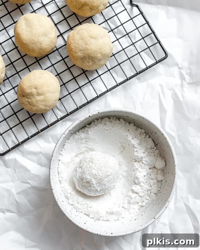 Flavorful Snowball Delights 7 Baked vegan snowball cookies alongside a bowl of powdered sugar, with some cookies already rolled and coated, showcasing the final step