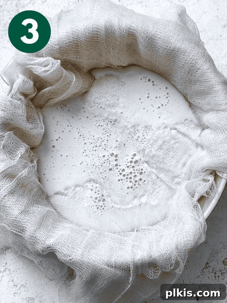 Process shot showing the blended oat mixture being strained through a nut milk bag into a bowl.