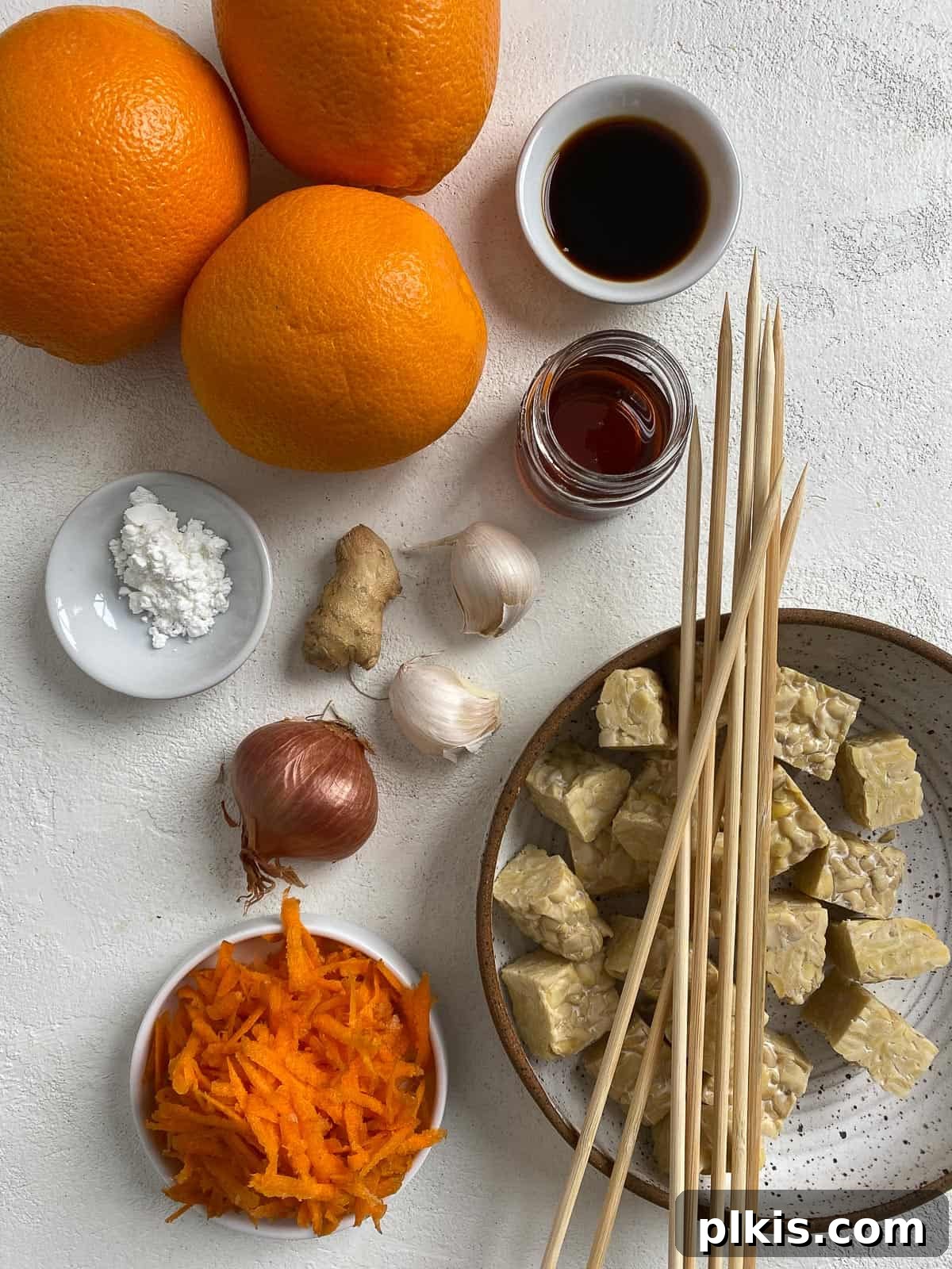 Fresh ingredients for Vegan Kabobs with Marinated Tempeh, including oranges, shallots, ginger, garlic, tempeh, and soy sauce, neatly arranged on a white surface.