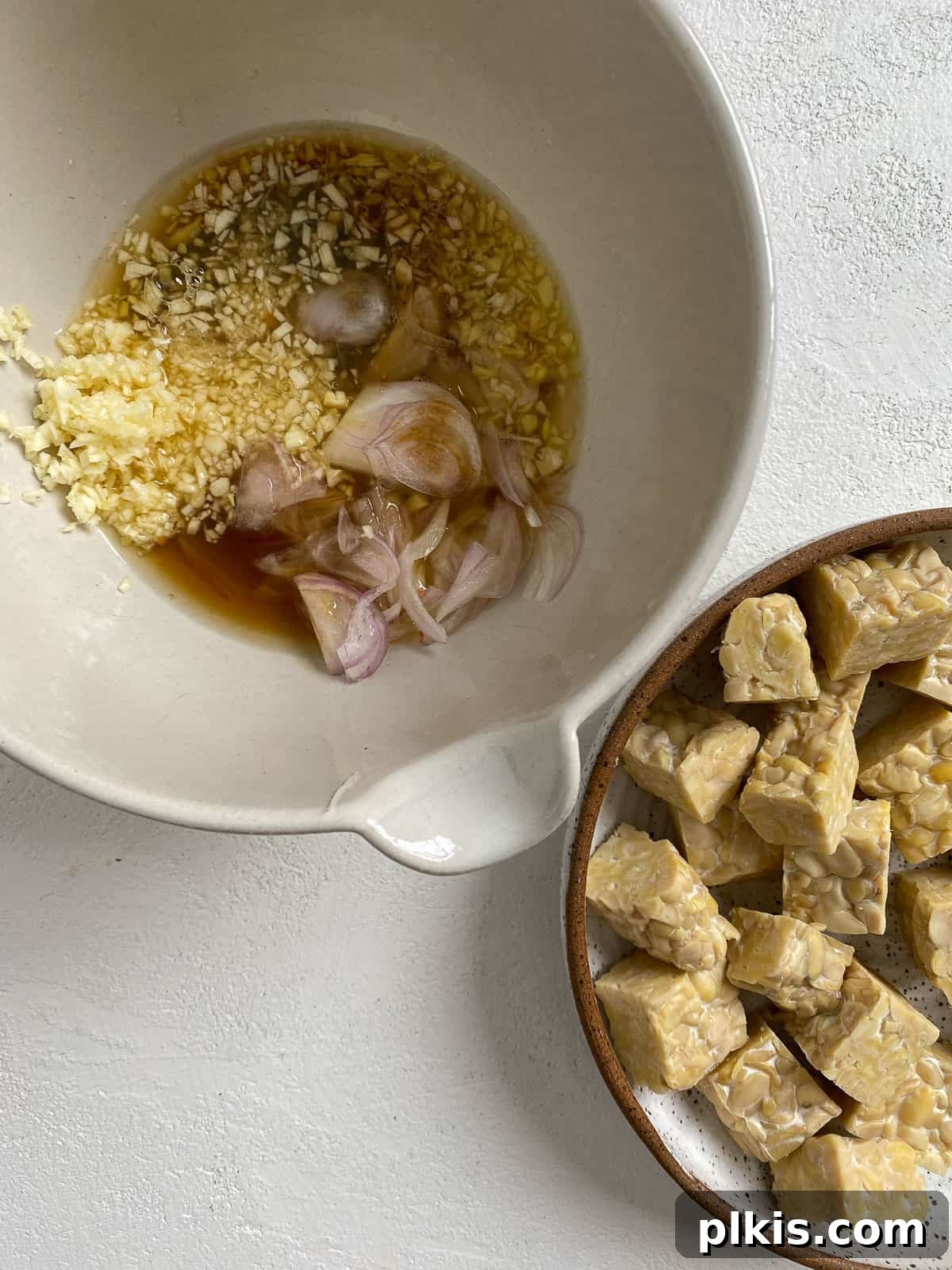 Marinating sauce in a bowl next to a bowl of cubed tempeh on a white surface, preparing for the marinade process.