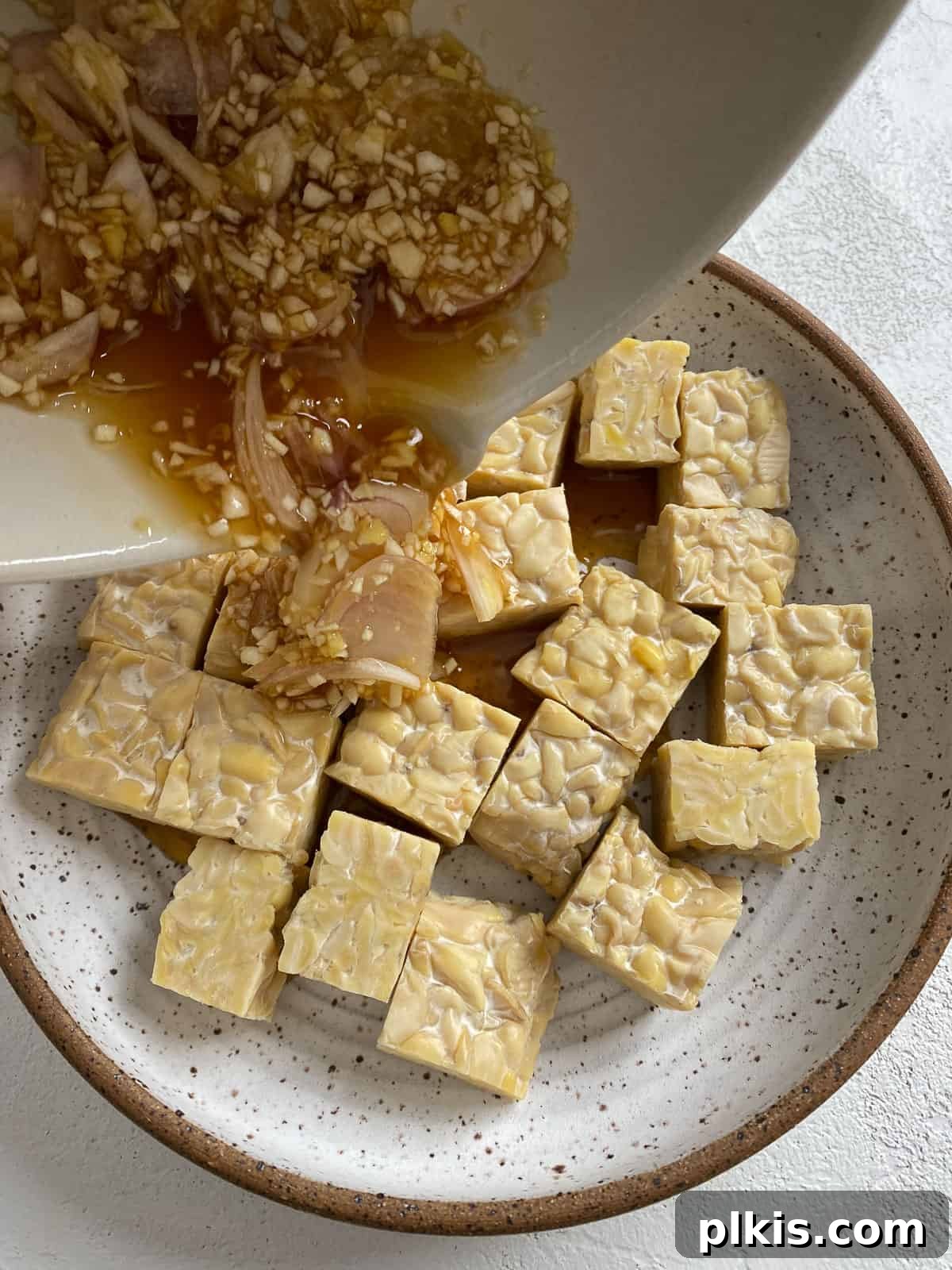 Process shot of pouring the freshly made marinade over cubed tempeh in a bowl, ensuring full coverage.