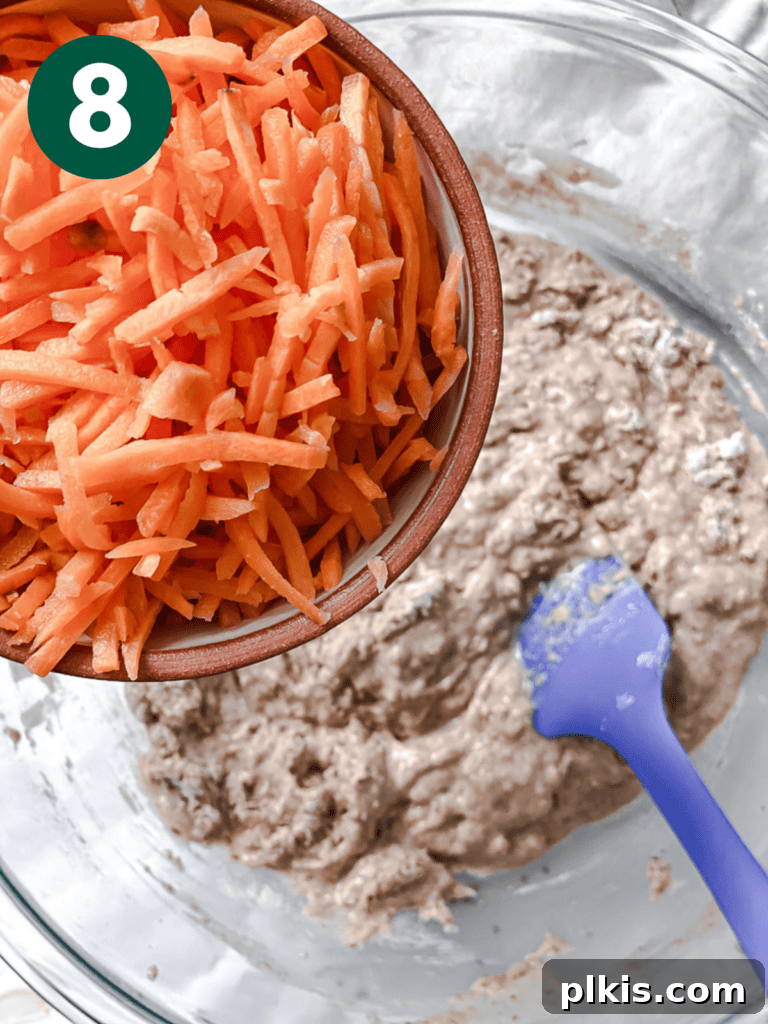 A process shot of shredded carrots being added to the cake batter in a mixing bowl.