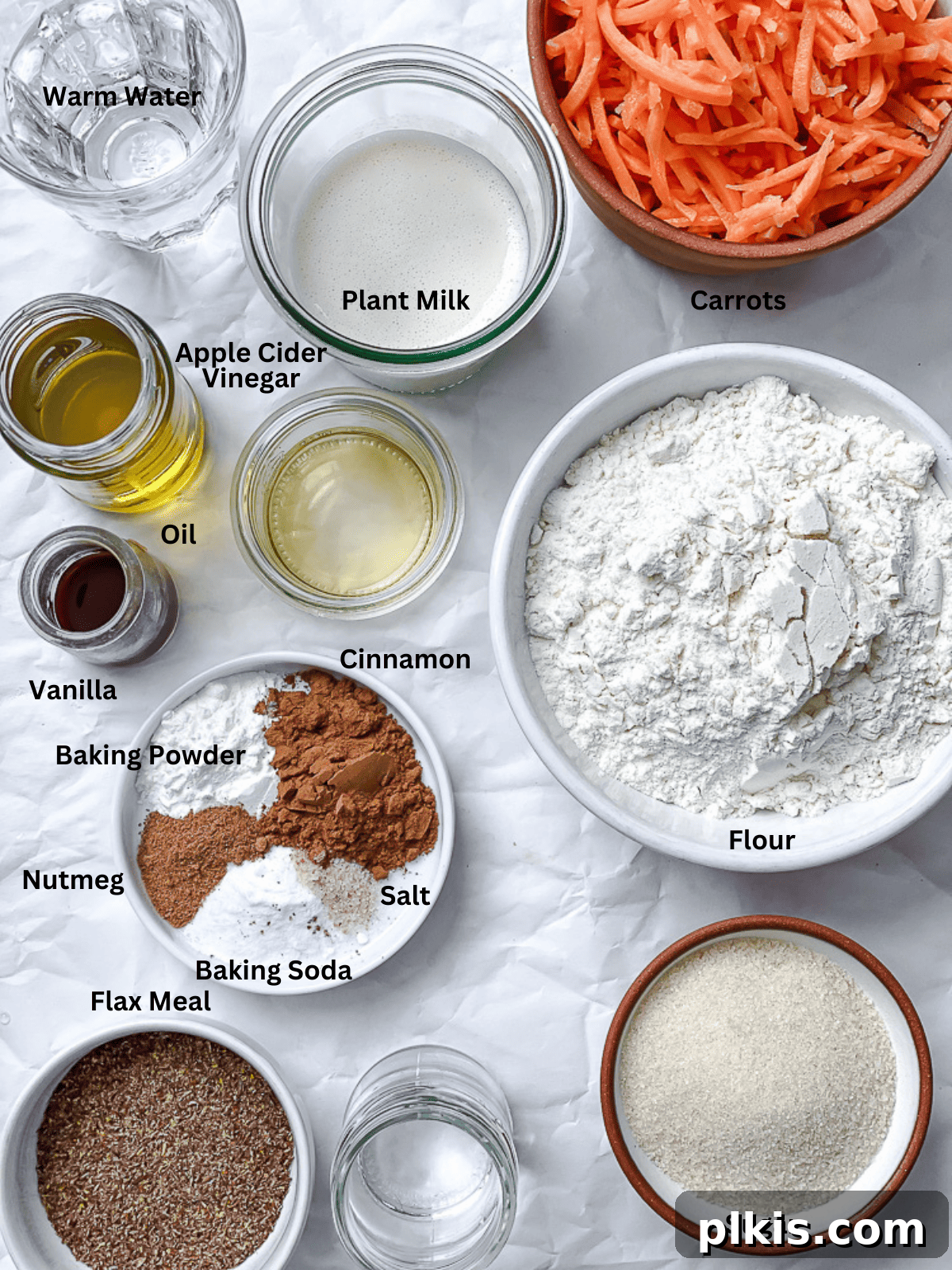 A vibrant display of fresh ingredients for Vegan Carrot Cake, including shredded carrots, walnuts, flour, sugar, and various liquids in bowls, all arranged on a white surface.
