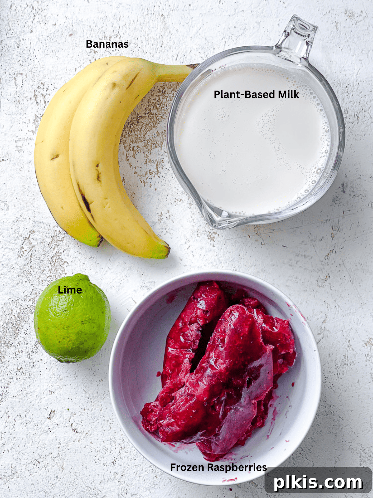 Effortless Raspberry Refresh 3 Individual ingredients for an Easy Raspberry Smoothie laid out on a white surface, including bananas, frozen raspberries, plant-based milk, and a lime.