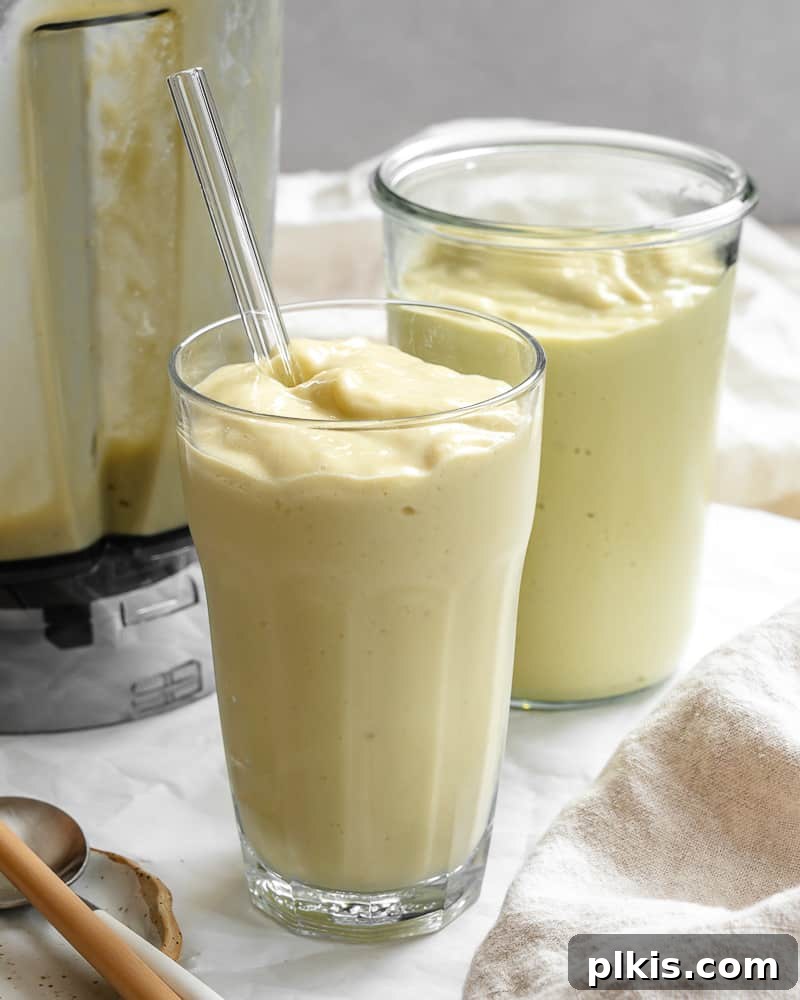 Tropical Mango Avocado Dream 2 Two glasses of creamy, yellow-green banana mango avocado smoothies, garnished with fresh mint leaves and mango cubes, on a light wooden surface.