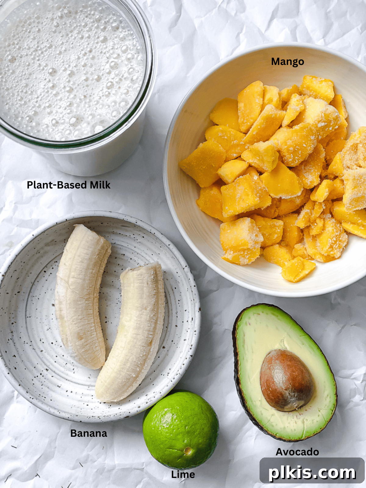 Tropical Mango Avocado Dream 3 Various ingredients for a mango avocado smoothie, including frozen mango chunks, a ripe banana, half an avocado, and a bottle of plant-based milk, arranged on a kitchen counter.