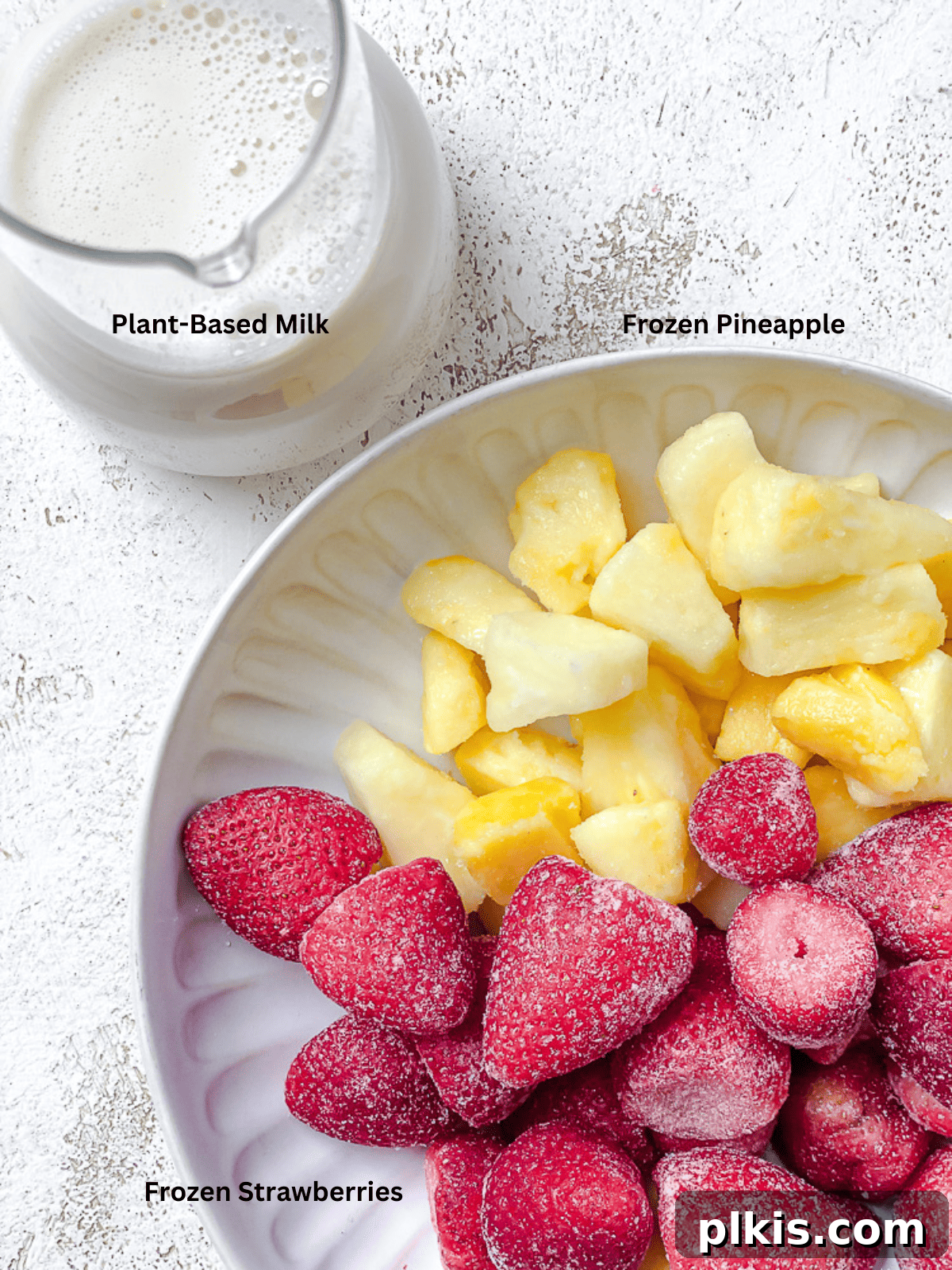 Fresh ingredients for Strawberry and Pineapple Smoothie laid out on a white surface, including strawberries, pineapple, and plant-based milk