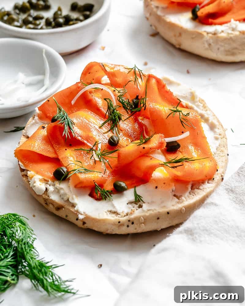 Smoky Carrot Lox 2 Completed Carrot Lox, showcasing thin, tender orange ribbons arranged artfully on a bagel half with vegan cream cheese, capers, and fresh dill, on a pristine white surface. This vibrant vegan smoked salmon alternative is ready for a delicious brunch or lunch.
