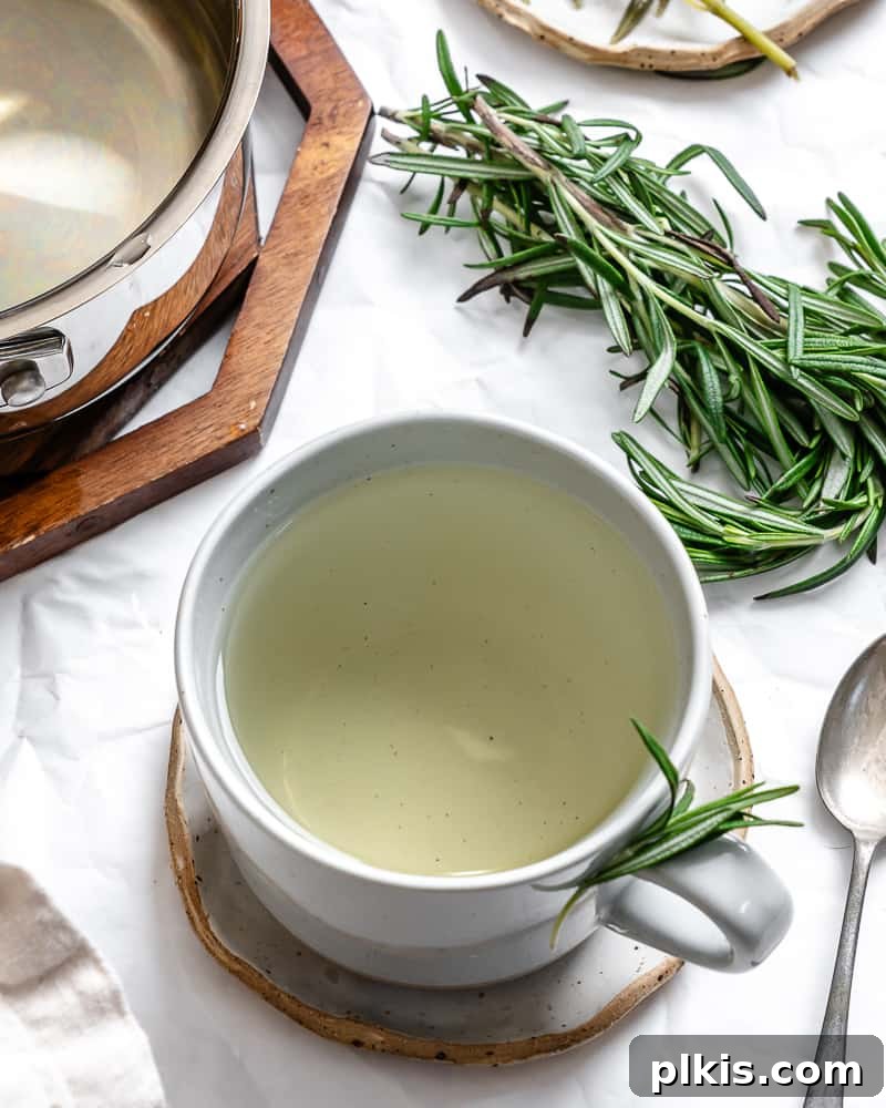 Rosemary's Evergreen Elixir 10 A white ceramic mug filled with golden rosemary tea, garnished with a fresh sprig of rosemary and resting on a rustic wooden surface.