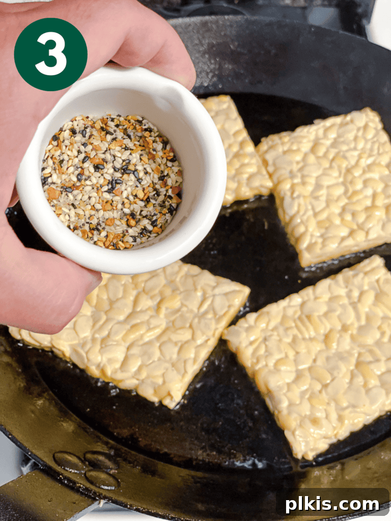 Savory Tempeh Sensation 6 Process shot showing everything bagel seasoning being sprinkled onto tempeh slices in a hot skillet.
