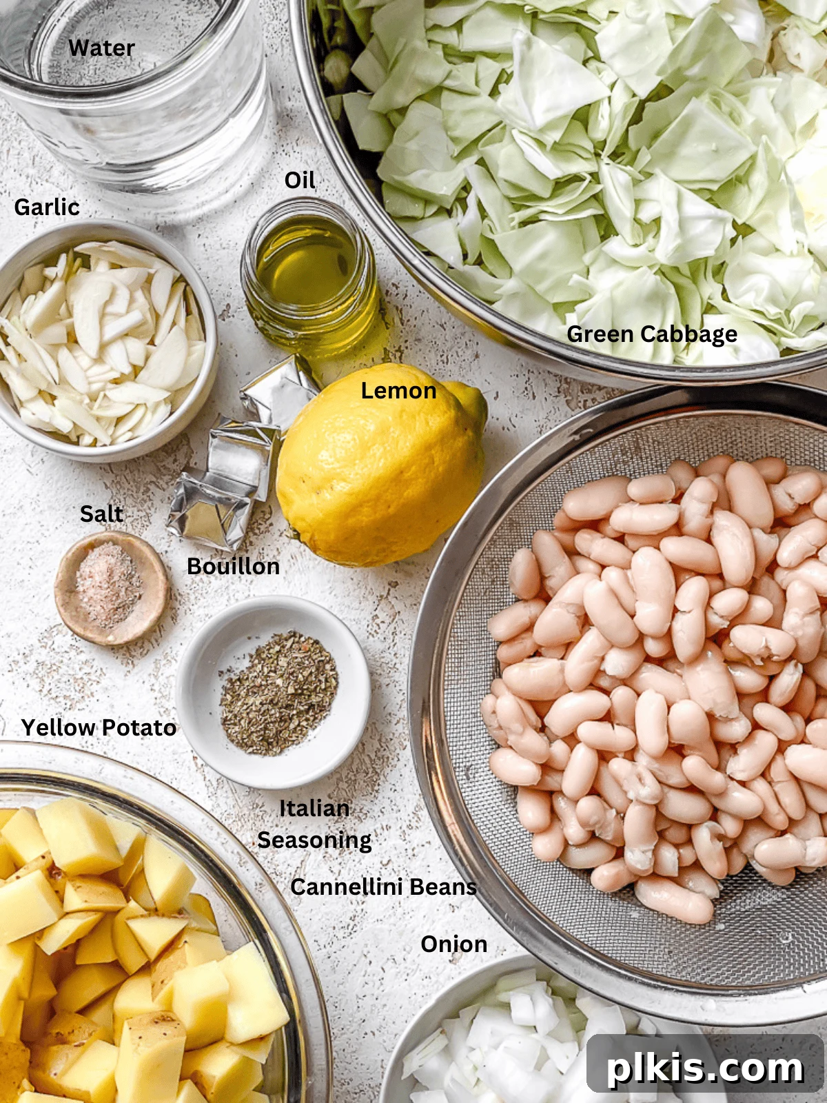 Rustic Cabbage and Bean Pot 3 Assortment of fresh ingredients for Cabbage Bean Soup laid out on a white surface, including cabbage, potatoes, onions, garlic, and canned beans.