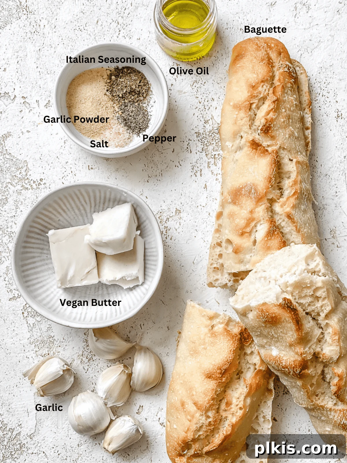 Effortless Plant-Based Garlic Bread 3 An overhead shot showcasing the fresh ingredients for easy vegan garlic bread laid out on a clean white surface: a baguette, fresh garlic cloves, a small bowl of garlic powder, olive oil, vegan butter, Italian seasoning, salt, and black pepper.